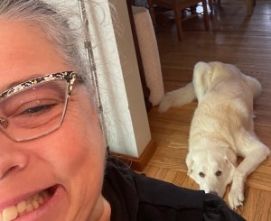 The author, wearing glasses and toothy grin. White dog laying down, but looking up behind me.